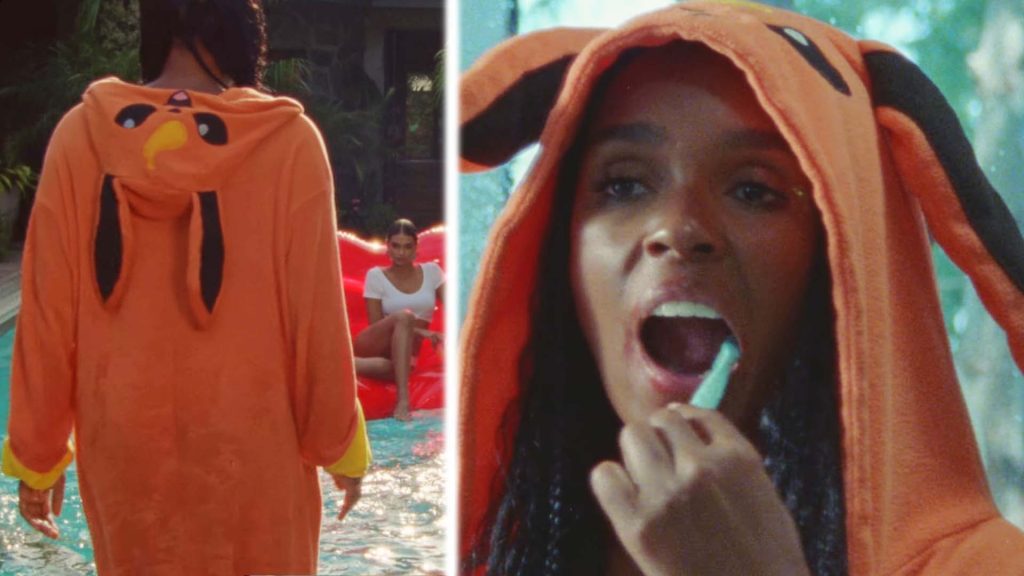 Janelle Monáe in a red dress and lipstick looking at her friend who is floating in a swimming pool with blue water and palm trees in the background.